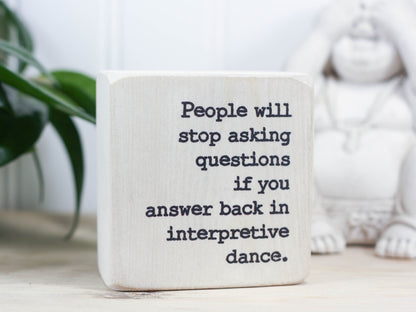 Small funny wood sign in whitewash with the saying "People will stop asking questions if you answer back in interpretive dance."