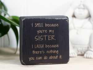 Small, freestanding, distressed black, solid wood sign with a funny saying on it "I smile because you're my sister. I laugh because there's nothing you can do about it."