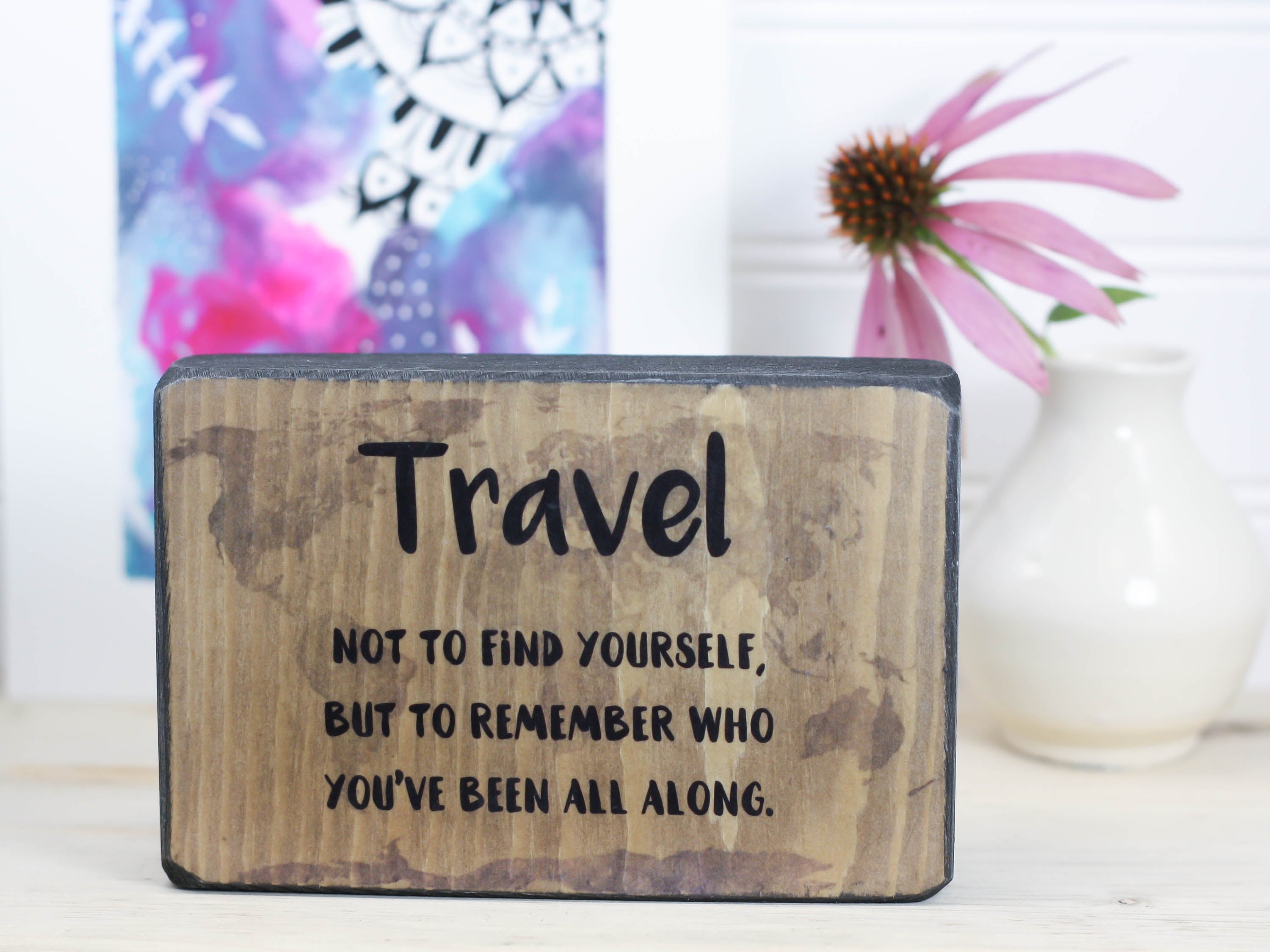 Small wood travel sign in distressed black with the saying "Travel not to find yourself, but to remember who you've been all along."
