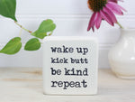 Small desk sign in whitewash with the saying "Wake up Kick butt Be kind Repeat".