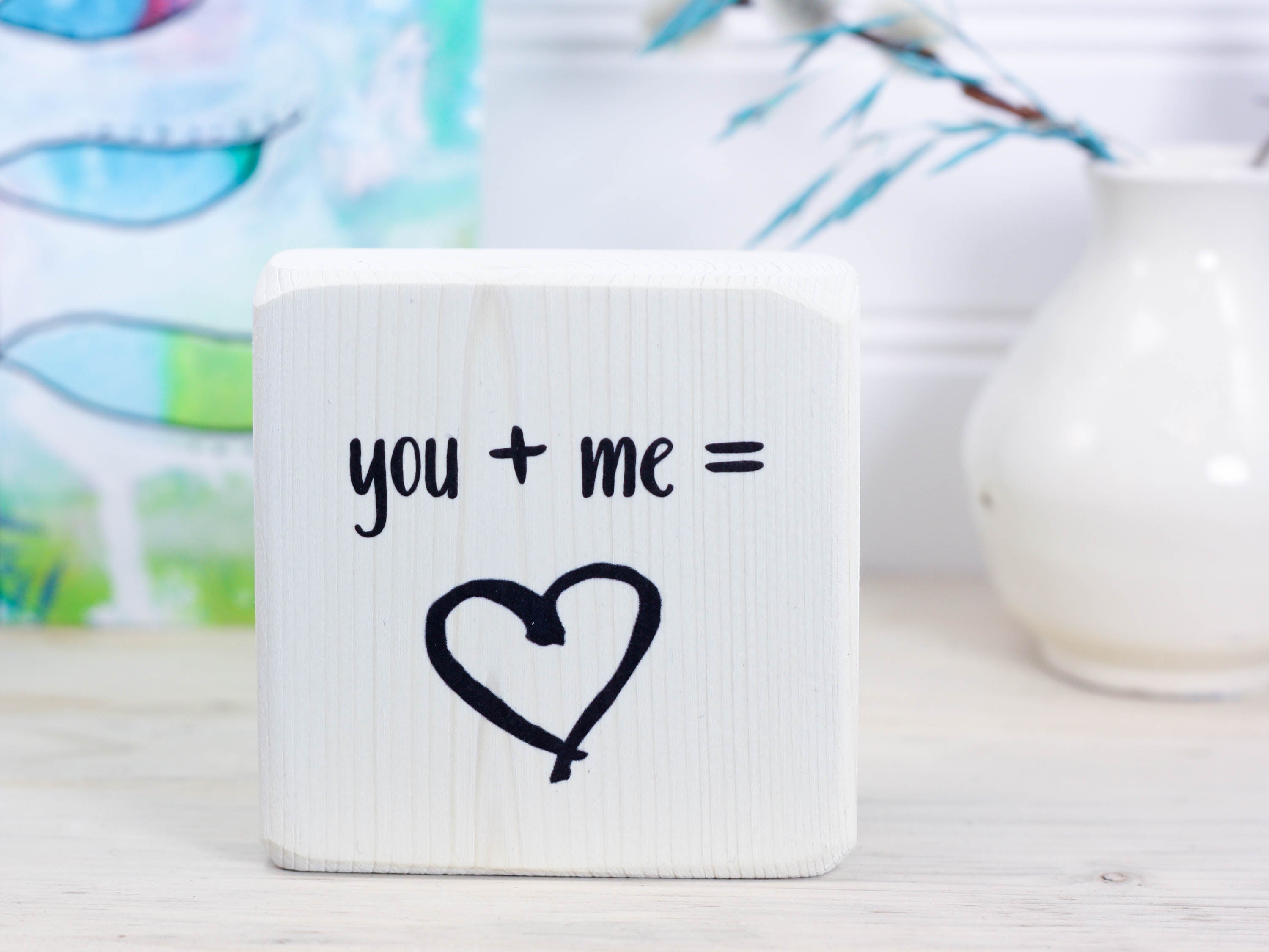 Mini wood shelf sign in whitewash with the text "you + me = (heart image)"