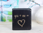 Mini wood desk sign in distressed black with the text "you + me = (heart image)"