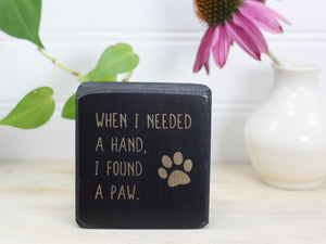 Small wood sign in distressed black with the saying "When I needed a hand, I found a paw".