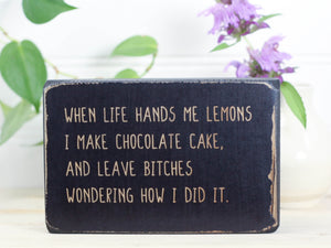 Small free-standing wood sign in distressed black with the saying "When life hands me lemons I make chocolate cake, and leave bitches wondering how I did it.